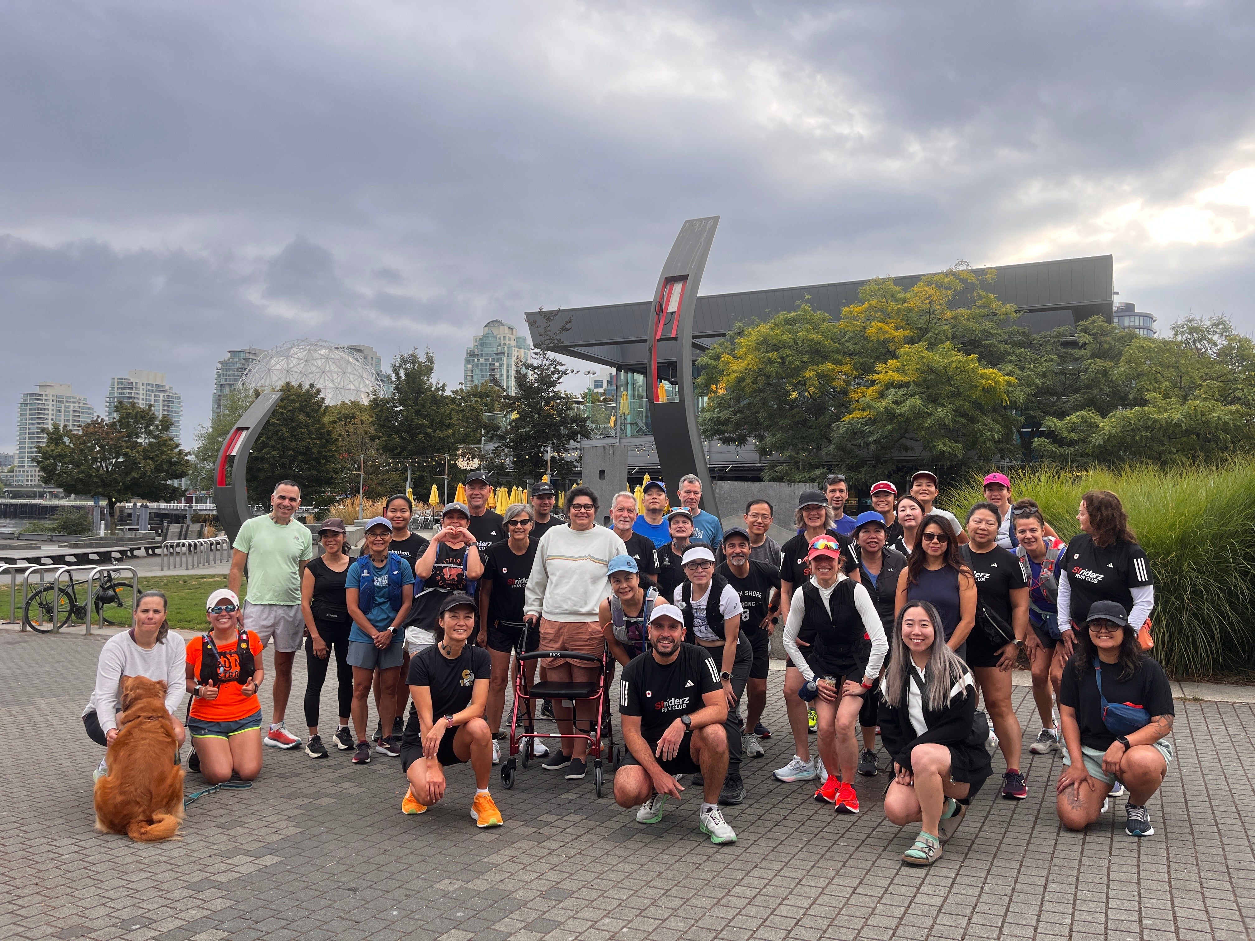 Group of Striderz run club runners posing for a photo outdoors with a cloudy sky and modern architecture in the background.
